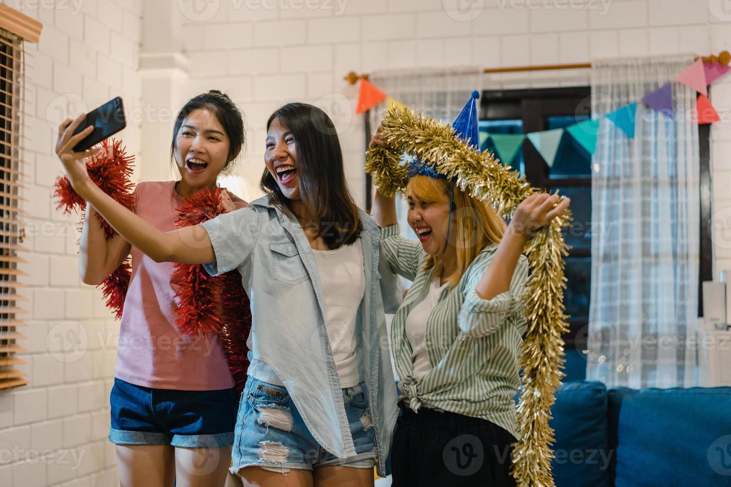 Group of Asian women party at home, female using mobile phone making vlog and selfie to social media while funny moment dancing together in living room in night. Young friend celebrate holiday concept photo