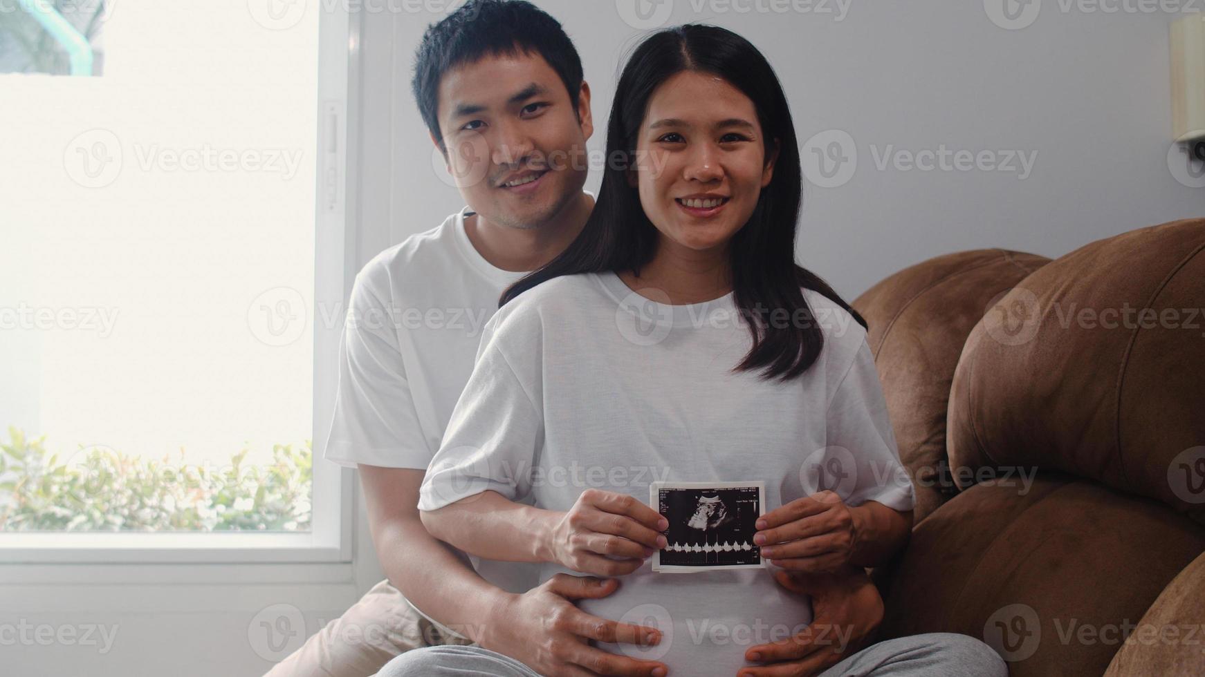 joven pareja asiática embarazada muestra y mira una foto de ultrasonido bebé en el vientre. mamá y papá se sienten felices sonriendo en paz mientras cuidan al niño acostado en el sofá en la sala de estar en el concepto de casa.
