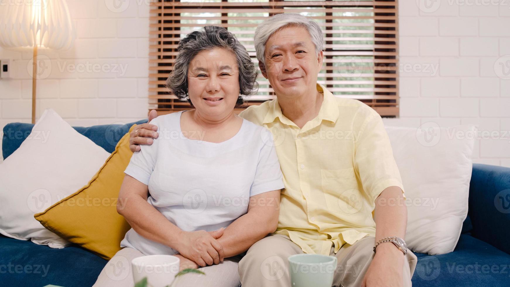 una pareja de ancianos asiáticos se siente feliz sonriendo y mirando a la cámara mientras se relaja en el sofá de la sala de estar en casa. Disfrutando del concepto de familia mayor en el hogar del estilo de vida del tiempo. retrato mirando a la cámara. foto