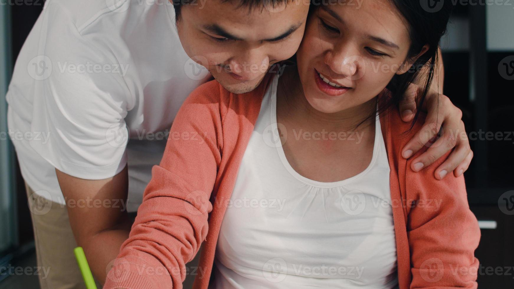 Young Asian Pregnant woman drawing baby in belly and family in notebook. Dad touch his wife belly while happy smiling positive and peaceful while take care child on table in living room at home. photo