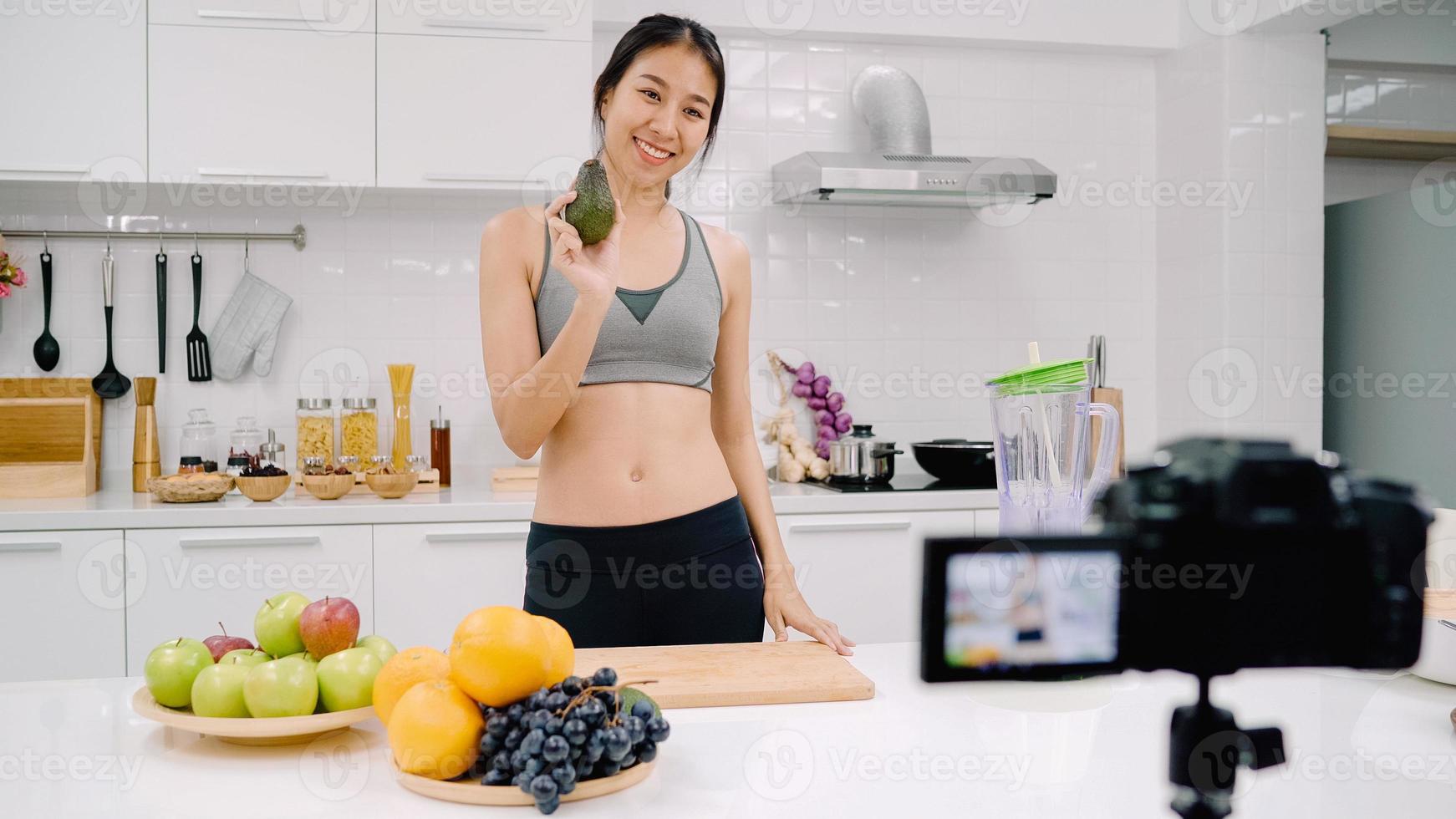 Blogger sporty Asian woman using camera recording how to make avocado juice video for her subscriber, female use organic fruit making avocado juice by herself at home. Healthy food concept. photo