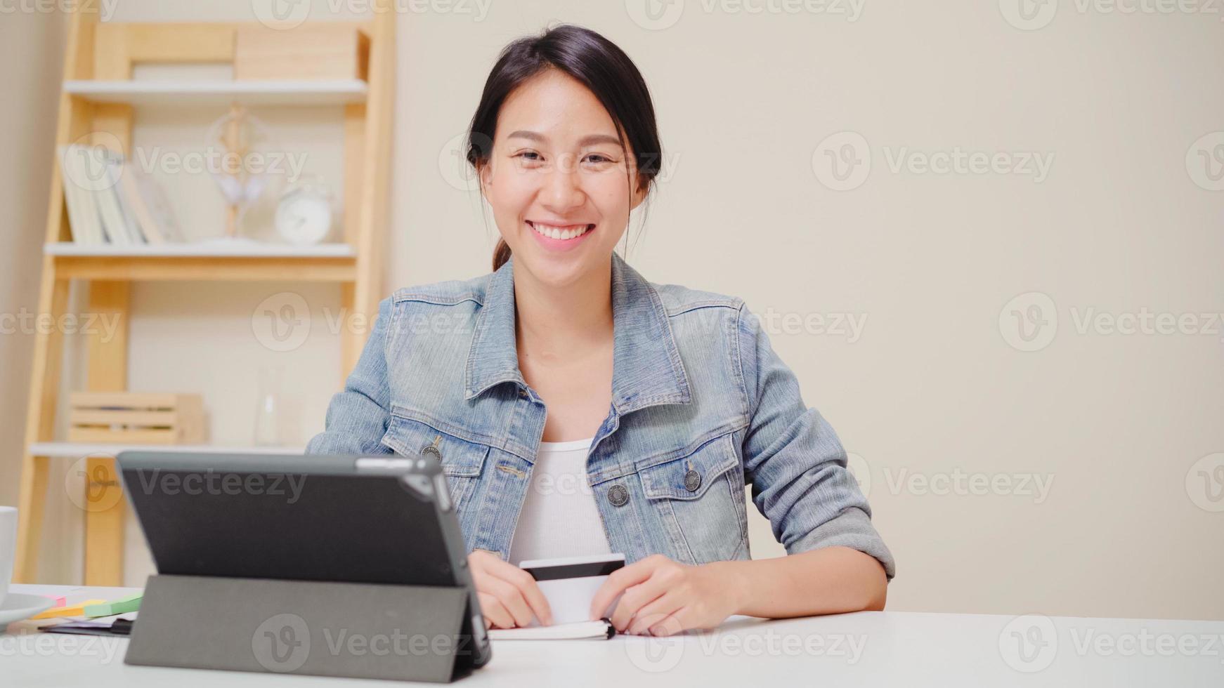 Beautiful smart business Asian woman using tablet buying online shopping by credit card while wear smart casual sitting on desk in living room at home. Lifestyle woman working at home concept. photo