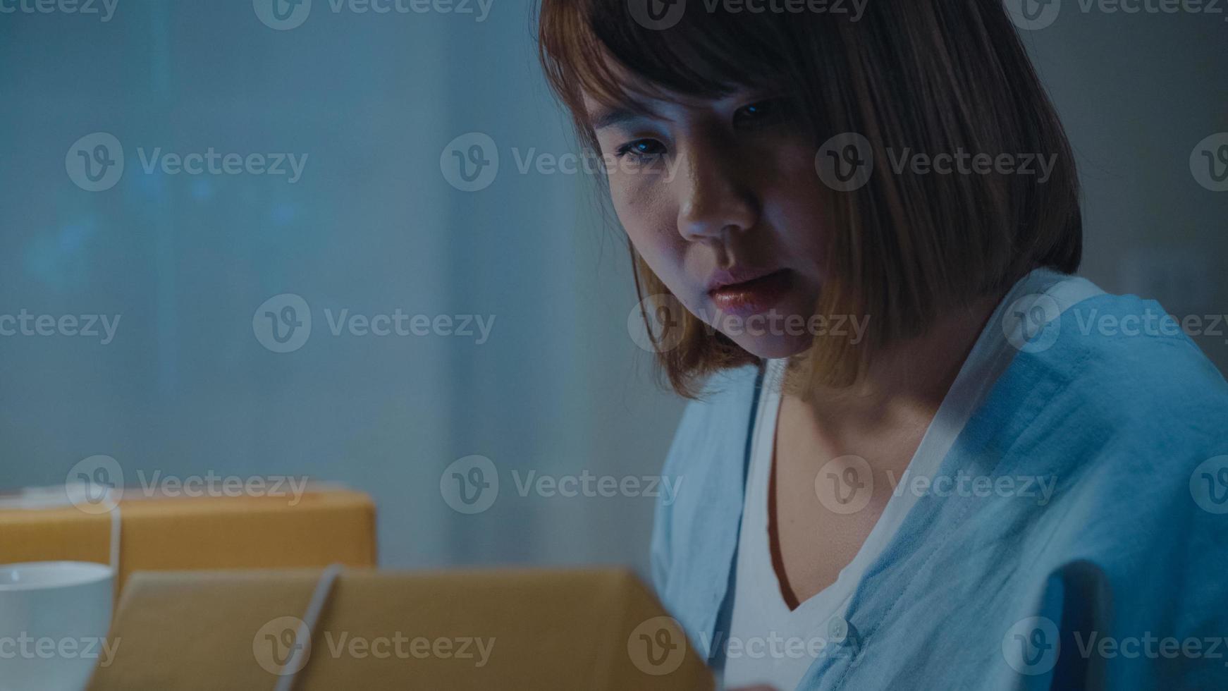 Beautiful smart Asian young entrepreneur business woman owner of SME online checking product on stock and save to tablet working late in night at home. Small business owner at home office concept. photo