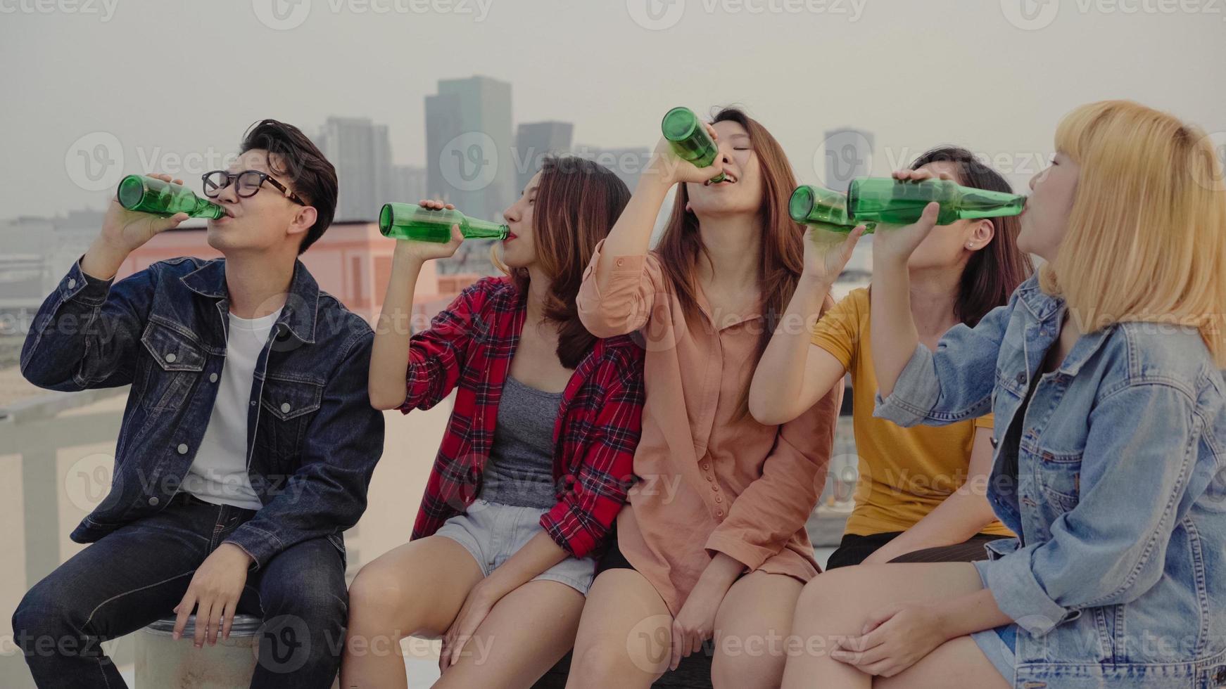 Group of young asian women and man people dancing and raising their arms up in air to the music played by dj at sunset urban party on rooftop. Young asian girls and boy friends hanging out with drinks photo