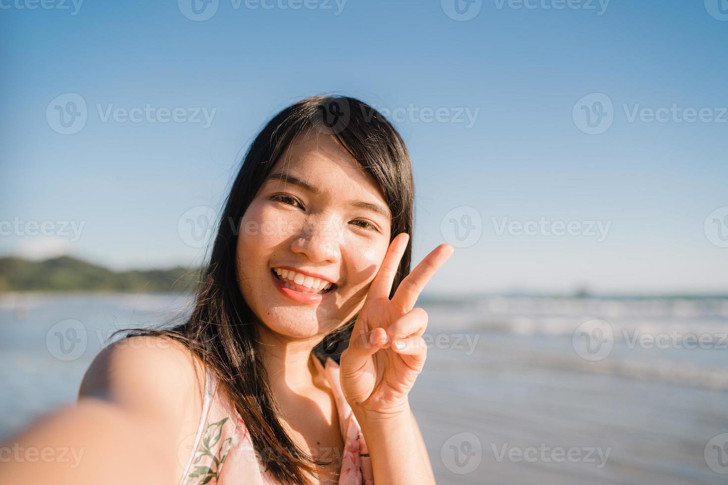 selfie de mujer asiática turística en la playa, joven hermosa mujer feliz sonriendo usando teléfono móvil tomando selfie en la playa cerca del mar cuando se pone el sol por la noche. las mujeres de estilo de vida viajan en concepto de playa. foto