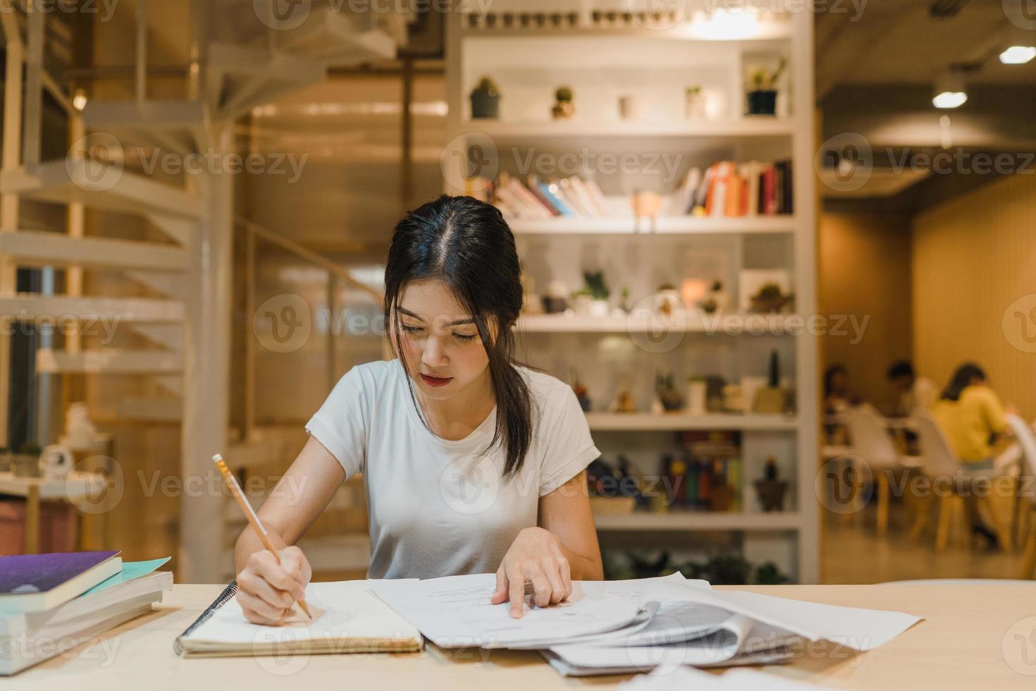 Asian student women reading books in library at university. Young undergraduate girl do homework, read textbook, study hard for knowledge on lecture desk at college campus overtime night. photo
