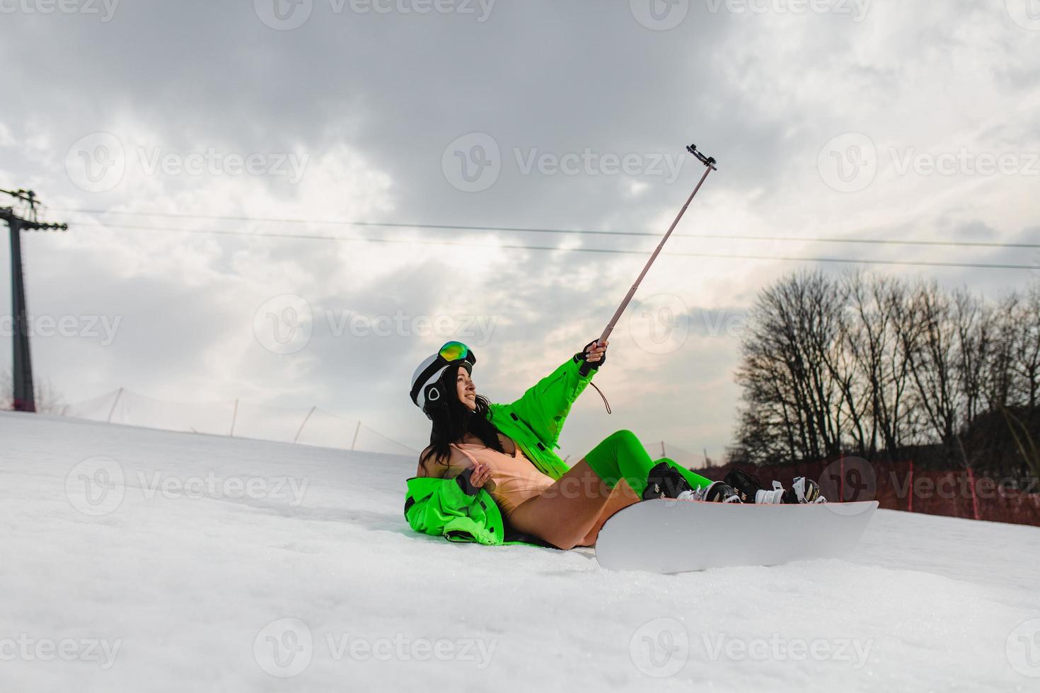 hermosa mujer usando un teléfono inteligente para hacer selfie con una tabla de snowboard en una pista de esquí foto