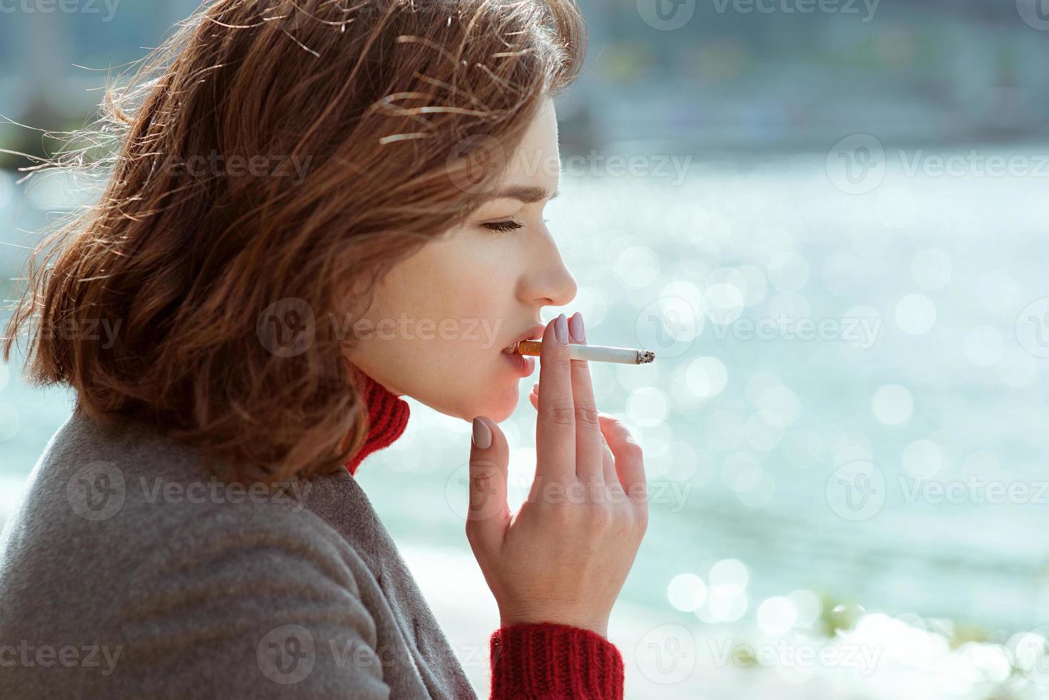 beautiful stylish sad stressful young woman in sweater and coat smoking cigarette near the river photo