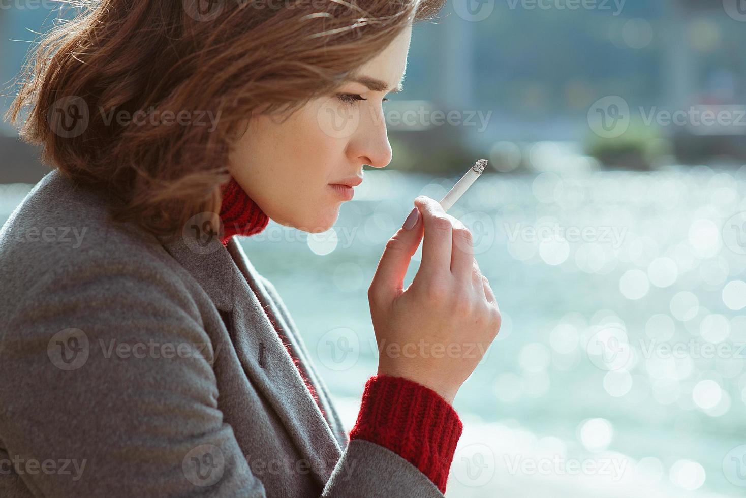 beautiful stylish sad stressful young woman in sweater and coat smoking cigarette near the river photo