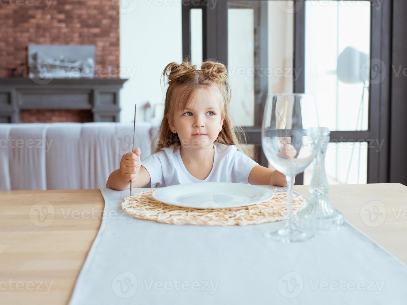 retrato de una adorable niña hambrienta con camiseta blanca sentada en la mesa con tenedor, cuchillo y plato vacío en el interior del loft foto