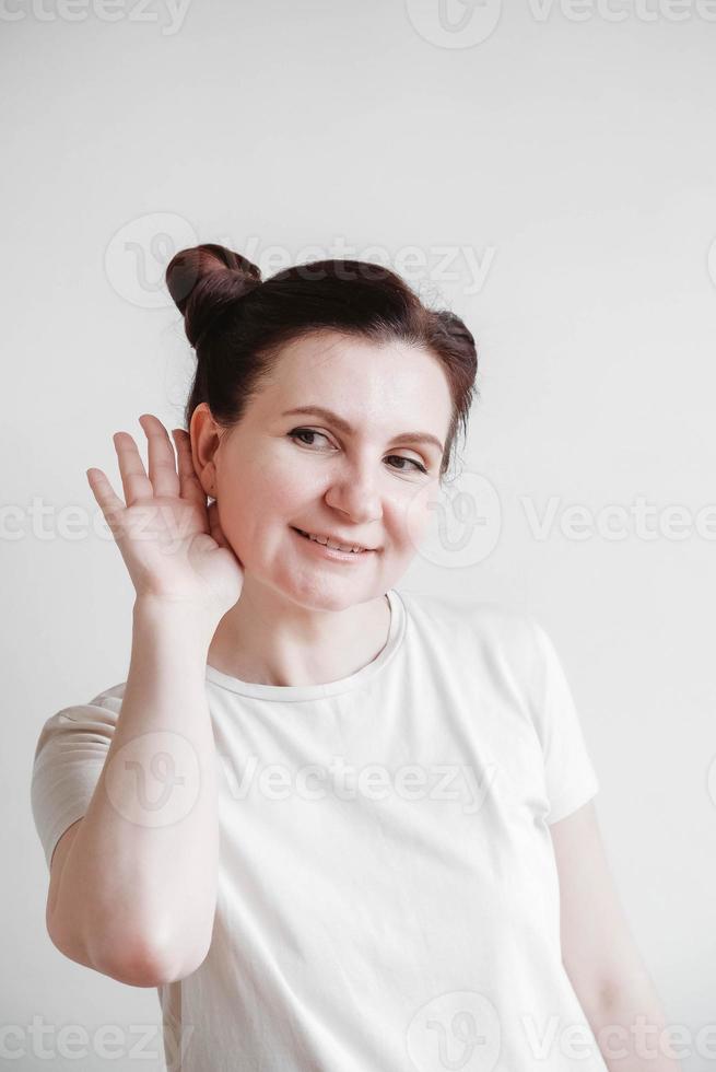 Woman raised her palm to her ear in a listening gesture on a white background. Copy, empty space photo