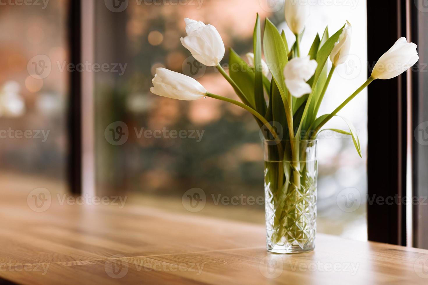 Vase with white flowers on big windows background with wooden table. Home coziness concept. Bouquet of tulips in glass vase. photo