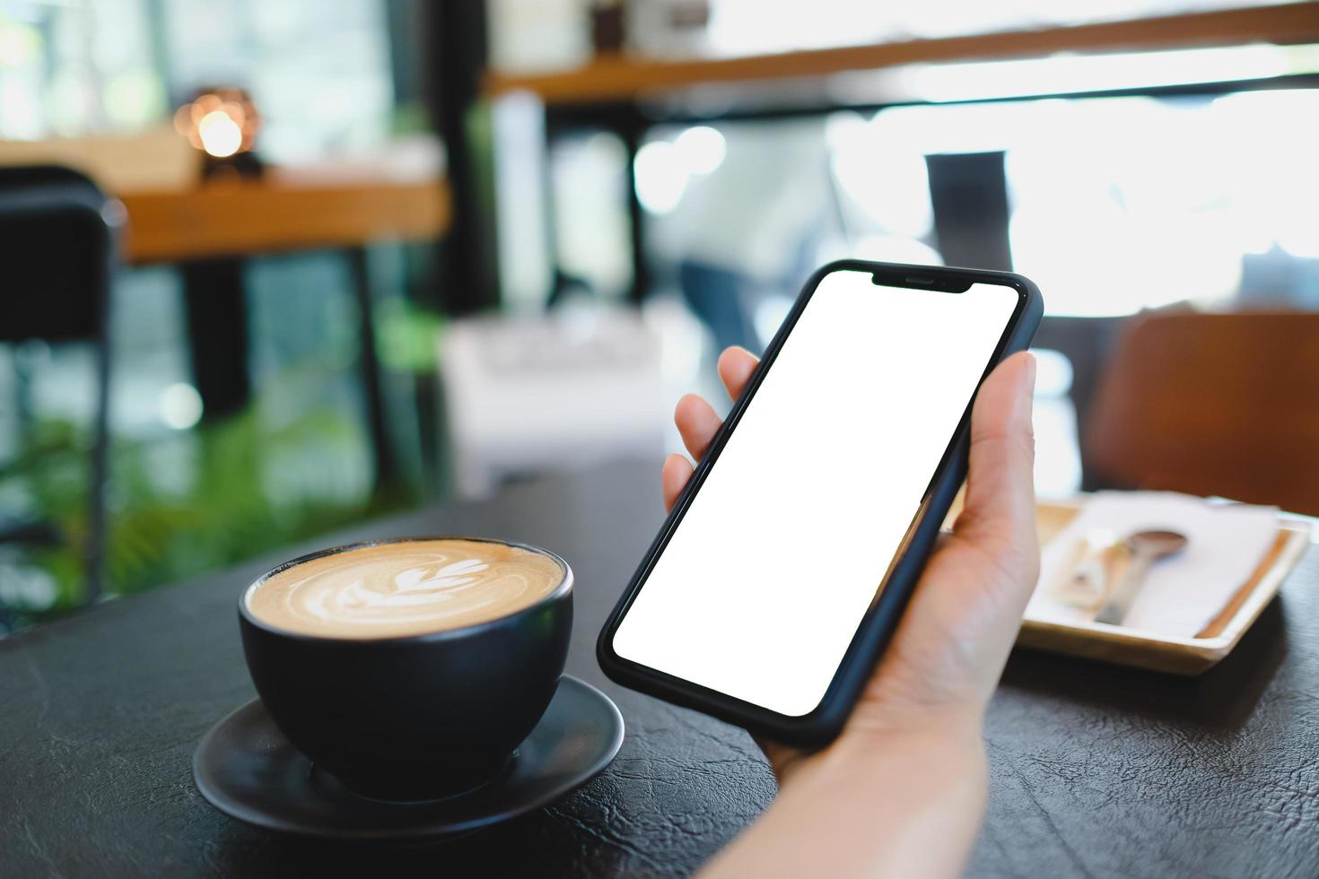 Mockup image of woman's hands holding white mobile phone with blank screen on thigh and coffee cup in cafe. photo
