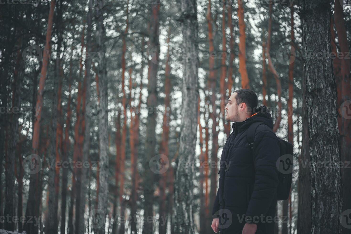 A young attractive man with a short beard and short hair in a black winter coat poses against the backdrop of a winter forest. photo