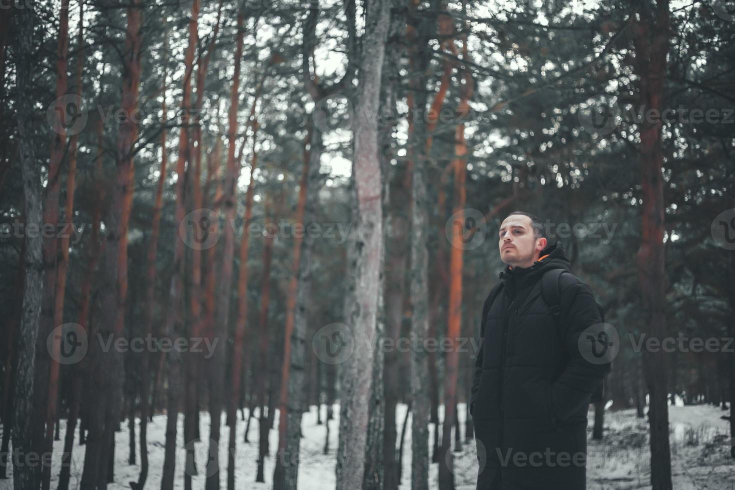 un joven atractivo con el pelo corto en un abrigo de invierno negro posa contra el telón de fondo de un bosque de invierno. foto