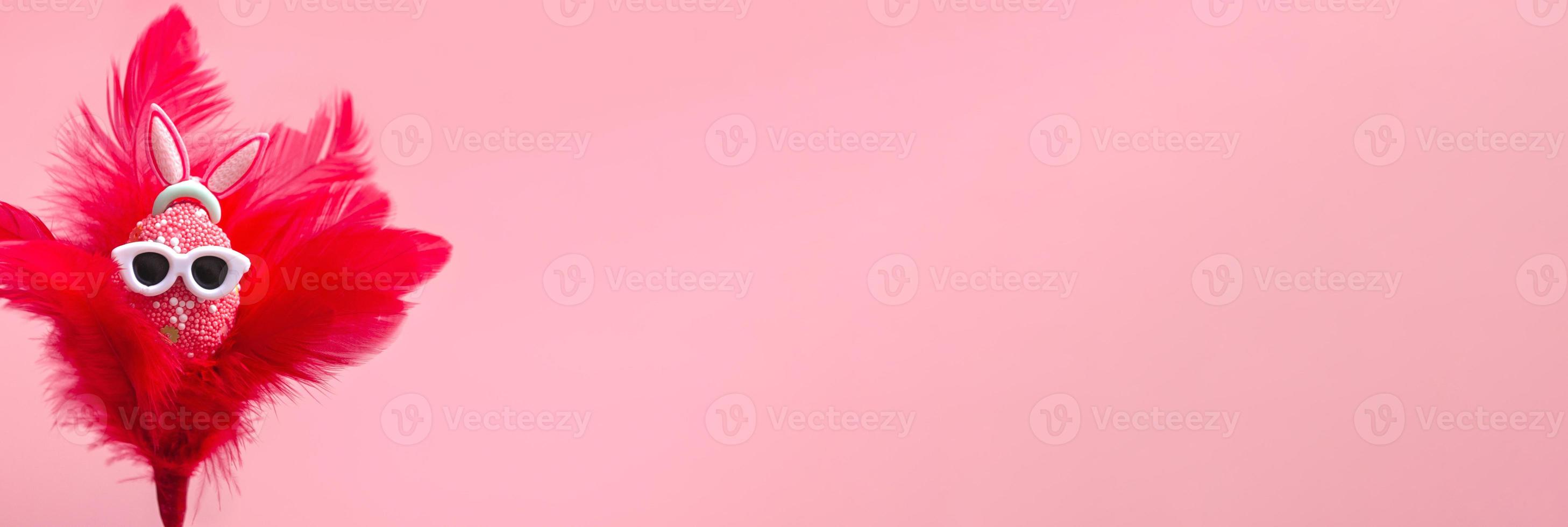huevo colorido con orejas de conejo en gafas de sol, en plumas rojas sobre un fondo rosa. banner de promoción de Pascua feliz, plantilla de maqueta. de cerca foto