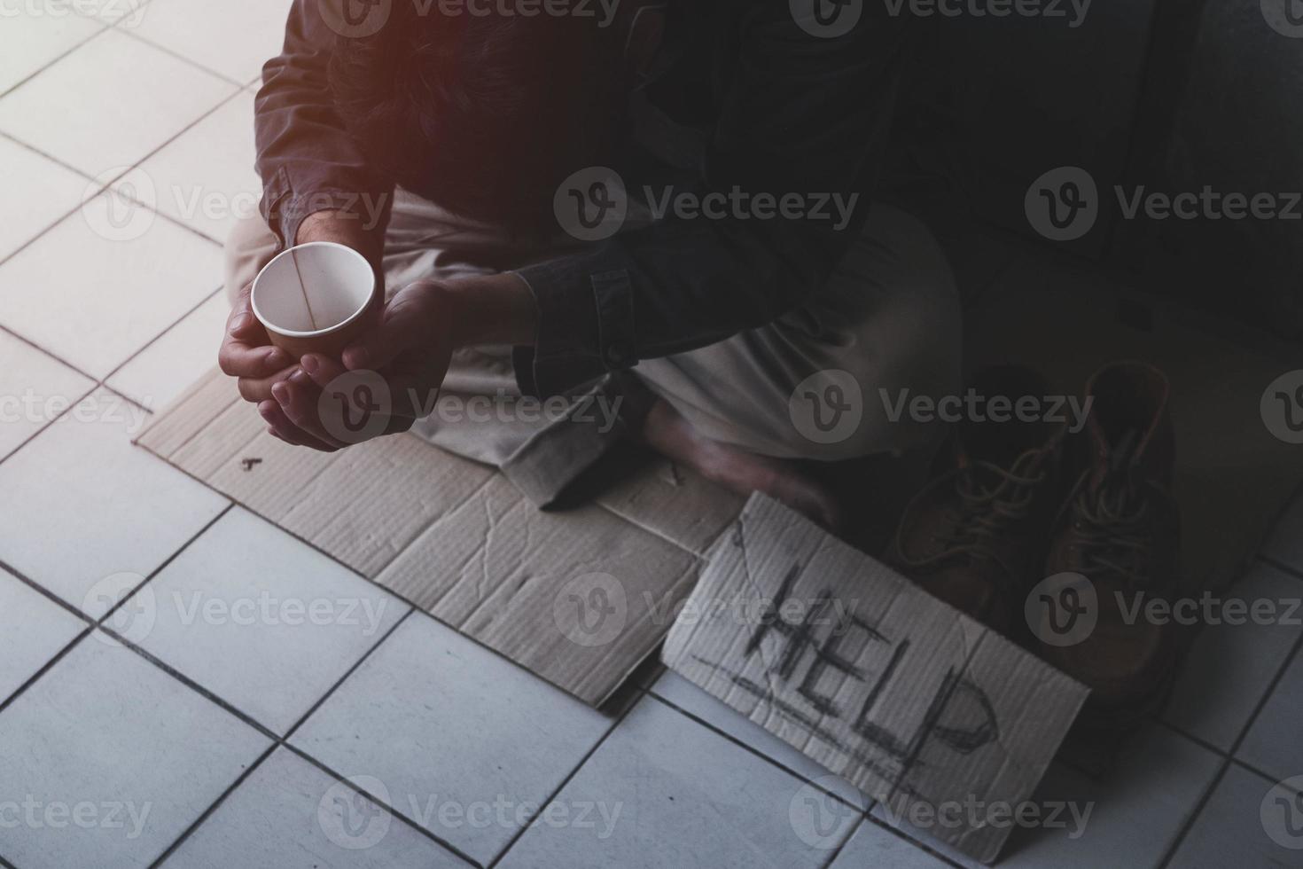 Homeless adult man sitting on the street in the shadow of the building and begging for help and money with sign.  Homeless concept. photo