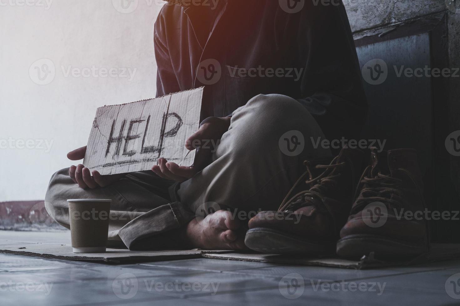 Homeless adult man sitting on the street in the shadow of the building and begging for help and money with sign.  Homeless concept. photo