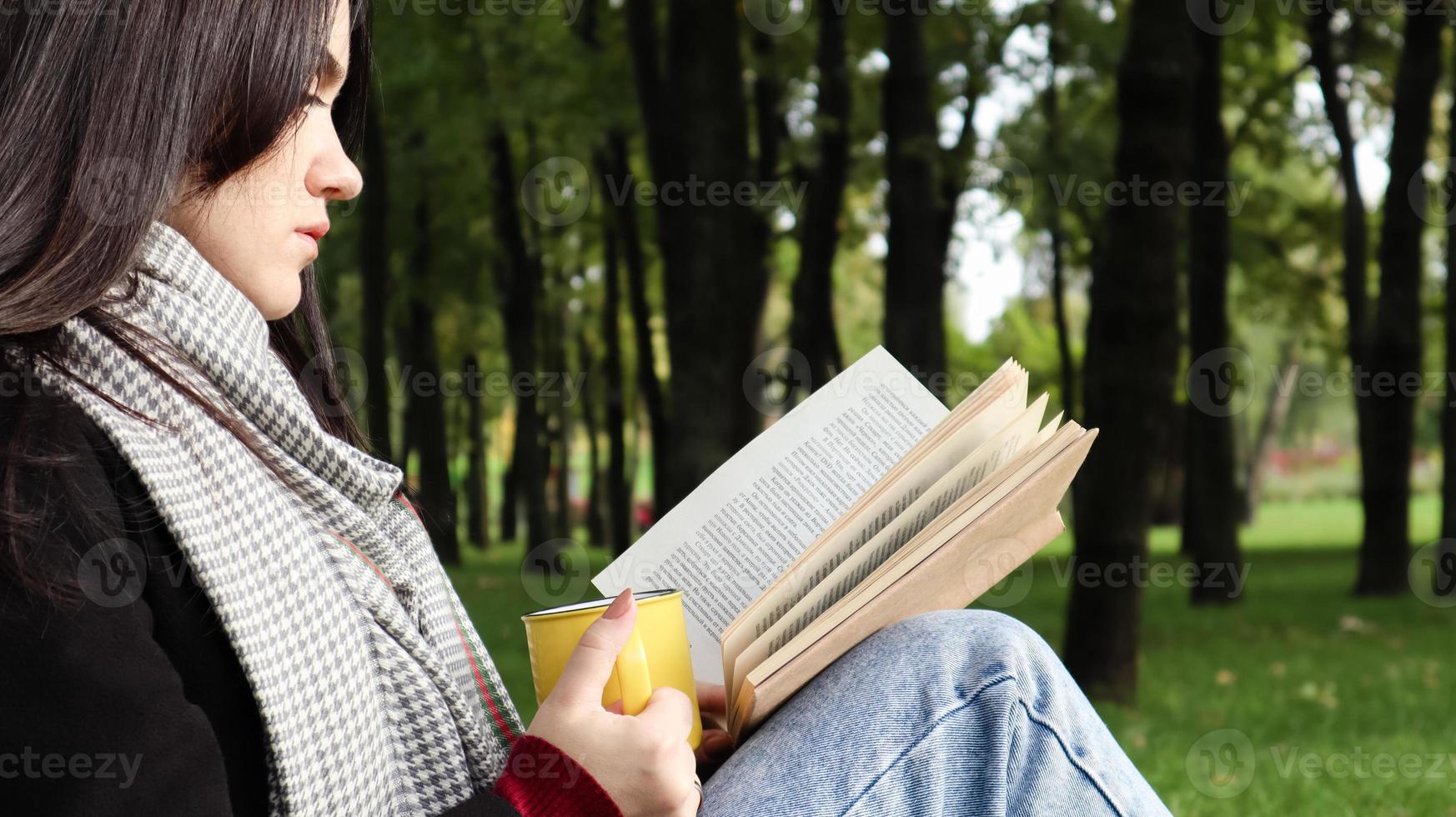 A woman sits under a tree and reads her favorite book while drinking coffee or tea from a yellow cup in a city park on green grass on a pleasant sunny day. Vacation, education and study concept. photo