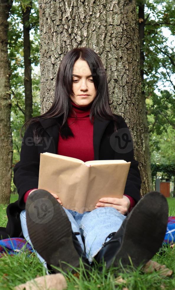Portrait of a beautiful young woman who is sitting under a tree and reading her favorite book in a city park on green grass on a pleasant sunny day. Vacation, education and study concept. photo