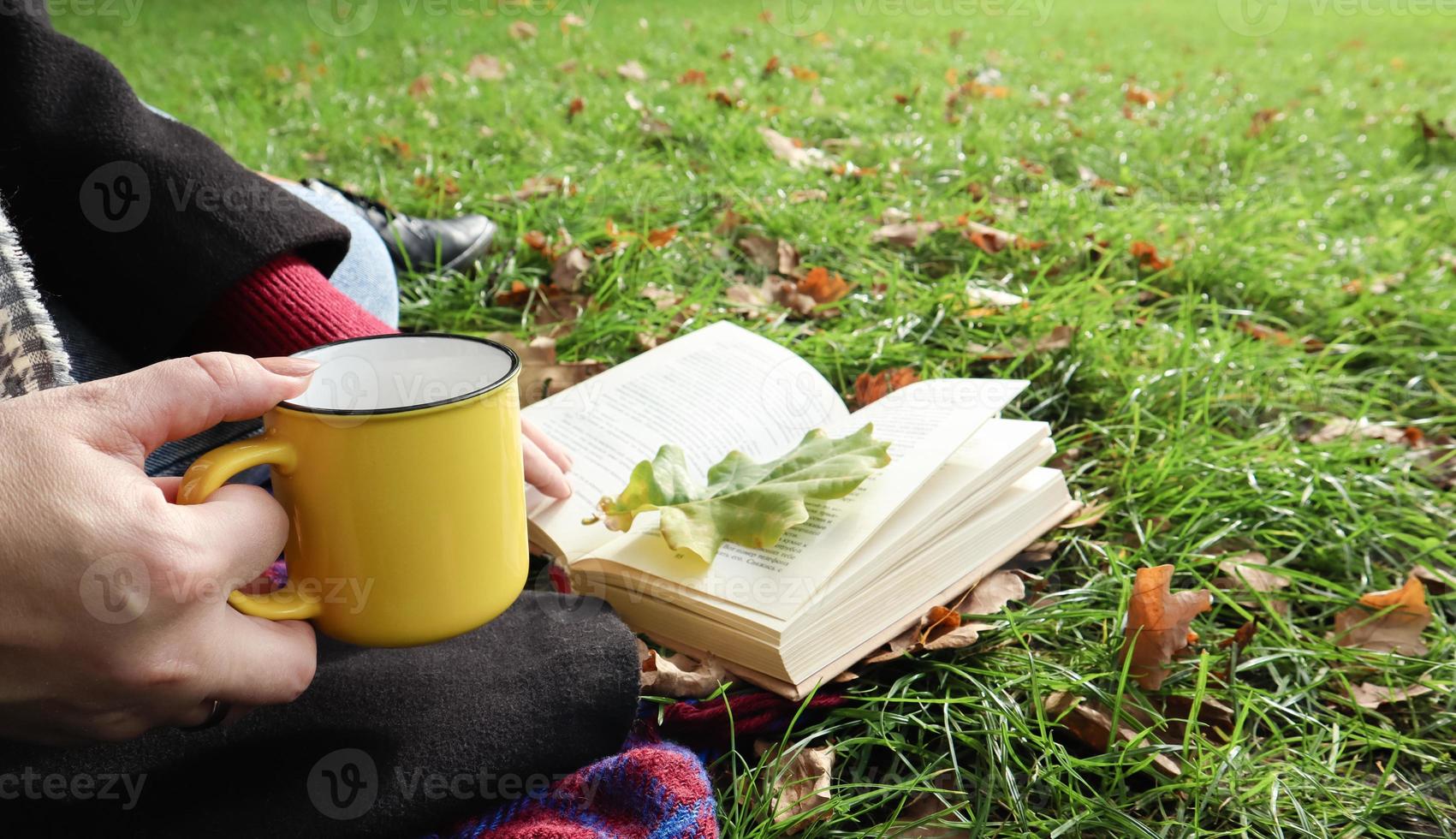 A girl sits on a blanket in the autumn forest, reads a book and drinks a hot drink from a yellow cup in a city park on a warm day on a green meadow. The concept of reading, relaxation and comfort. photo