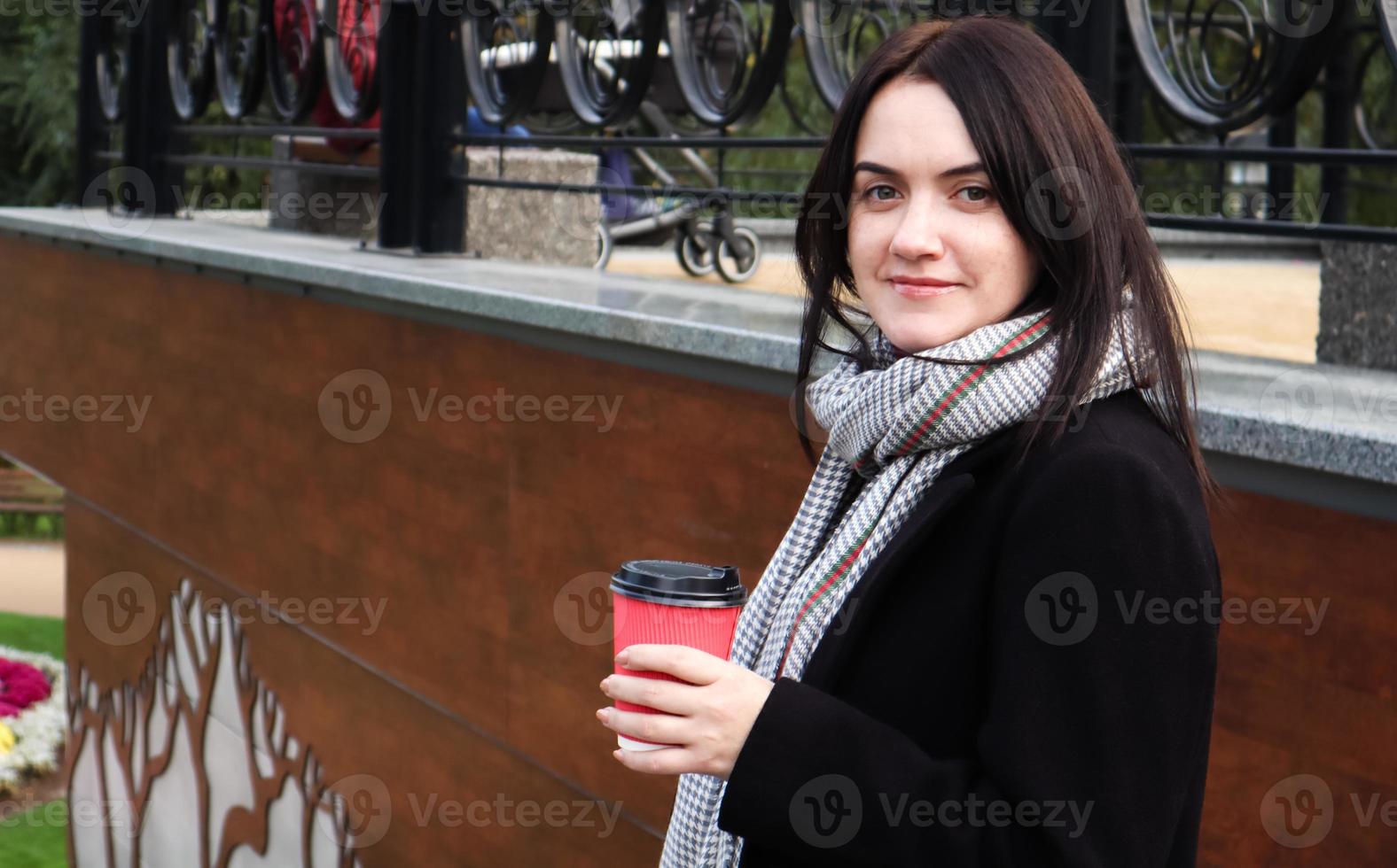 una joven mujer elegante con abrigo y bufanda bebe café caliente por la mañana en un vaso de papel ecológico rojo al aire libre en un parque de otoño. retrato, de, un, mujer joven, tenencia, un, taza de café para llevar, poca, profundidad de campo foto