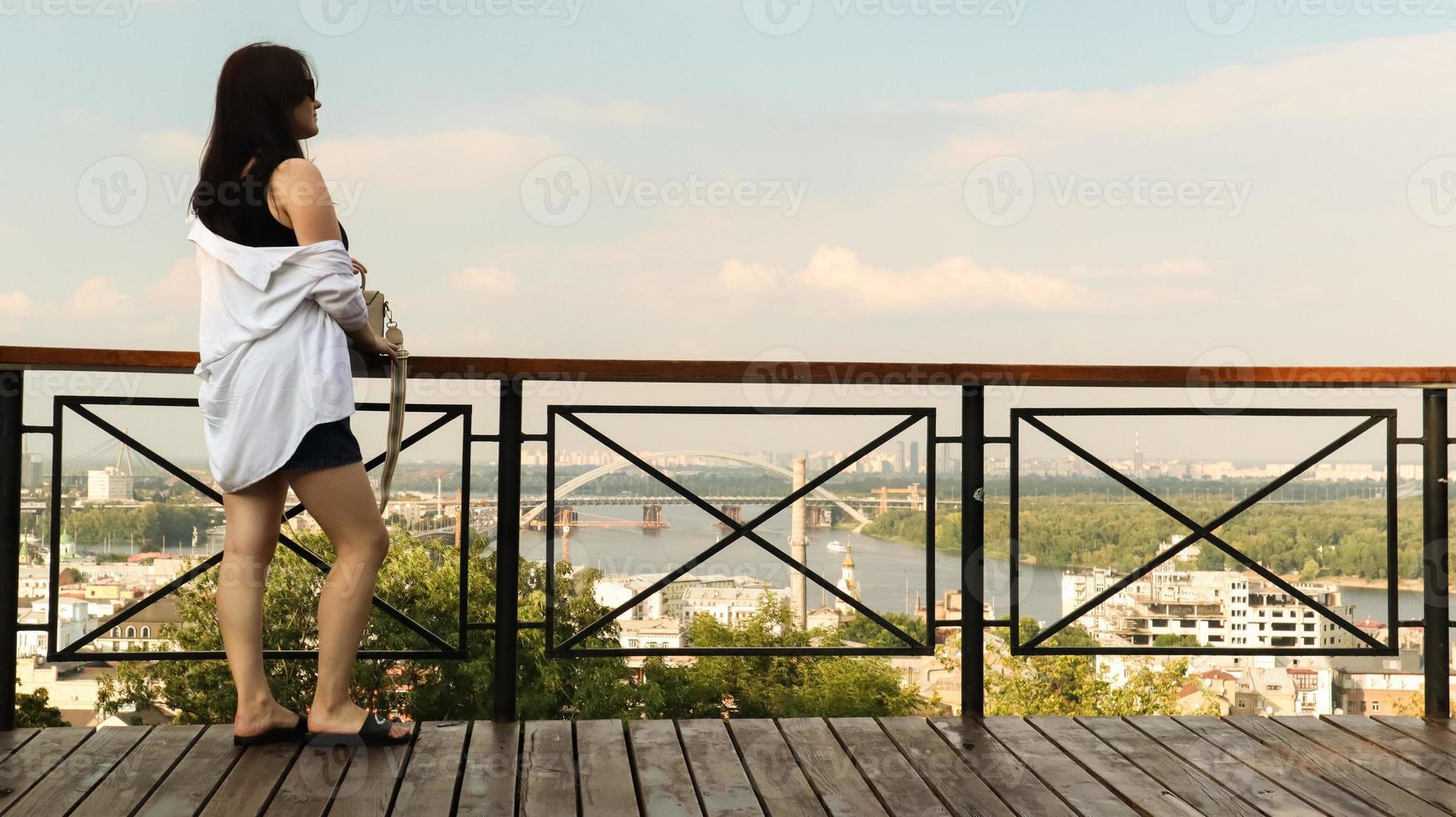 Back view of a young woman in a white shirt, a brunette traveler looks at the cityscape on a sunny day from a high hill with an observation deck for tourists. Vintage photo