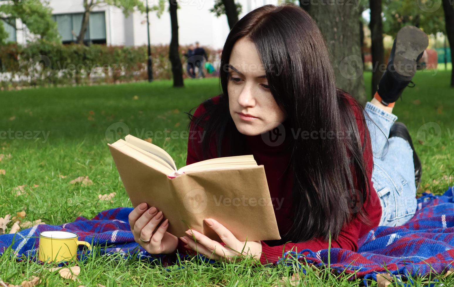 A young woman lies and reads her favorite book on a woolen plaid blanket or blanket in a city park on green grass on a pleasant sunny day. The concept of recreation and leisure, education and study. photo