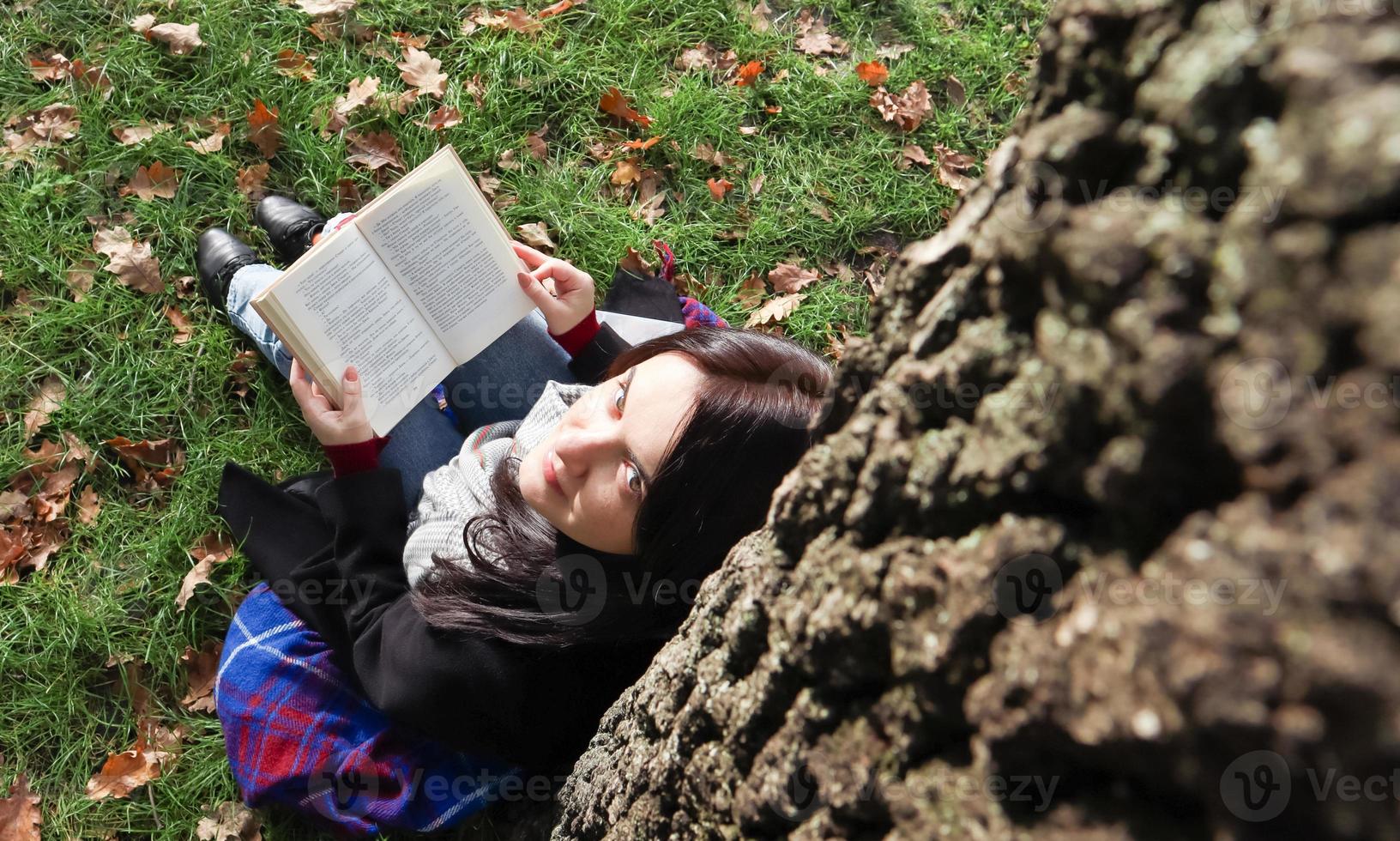 Portrait of a beautiful woman who sits under a tree and reads her favorite book in a city park on green grass on a pleasant sunny day. Vacation, education and study concept. View from above. photo