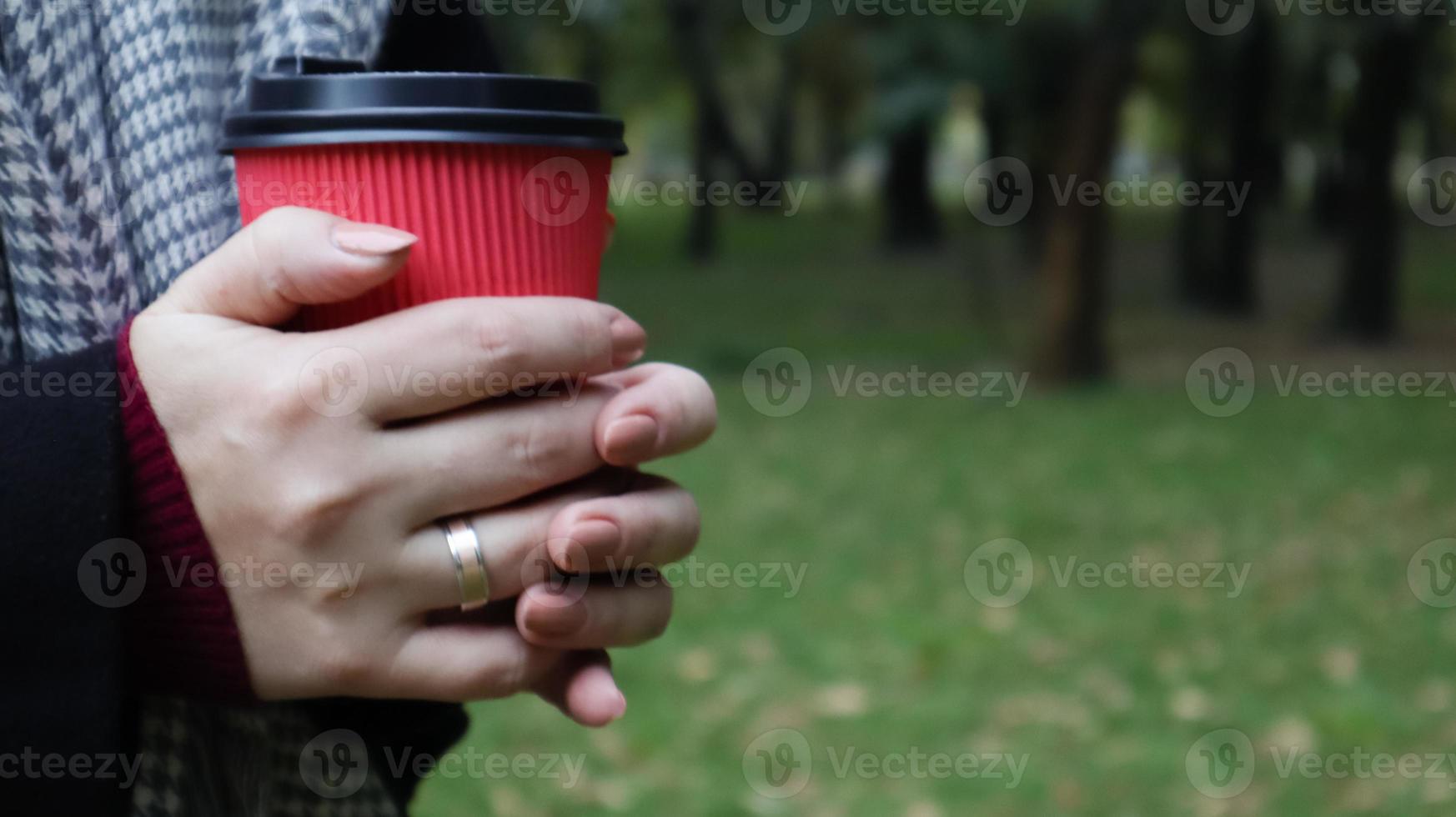 A young girl in a coat holds morning coffee with her while walking in the park. Hand holding paper cup of coffee in green park. Takeaway cappuccino. Close-up, copy space. photo