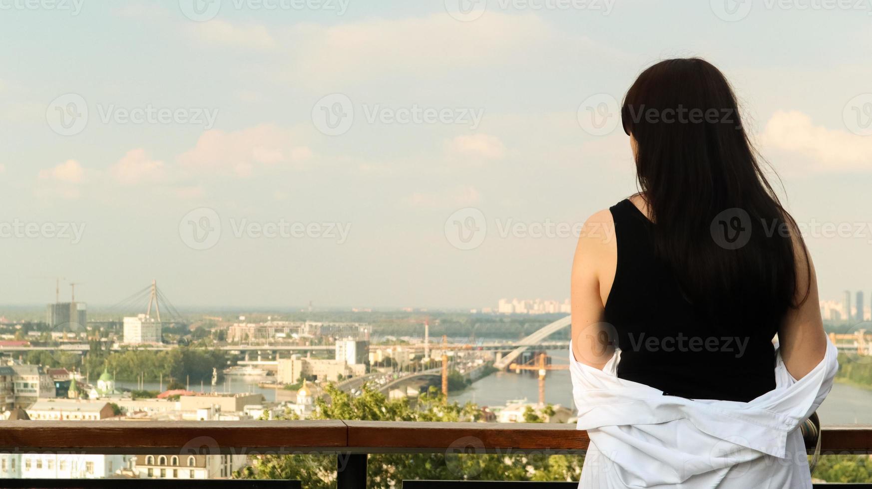 Back view of a young woman in a black T-shirt and white shirt, a brunette traveler looks at the cityscape on a summer sunny day from a high hill with an observation deck for tourists. Vintage photo