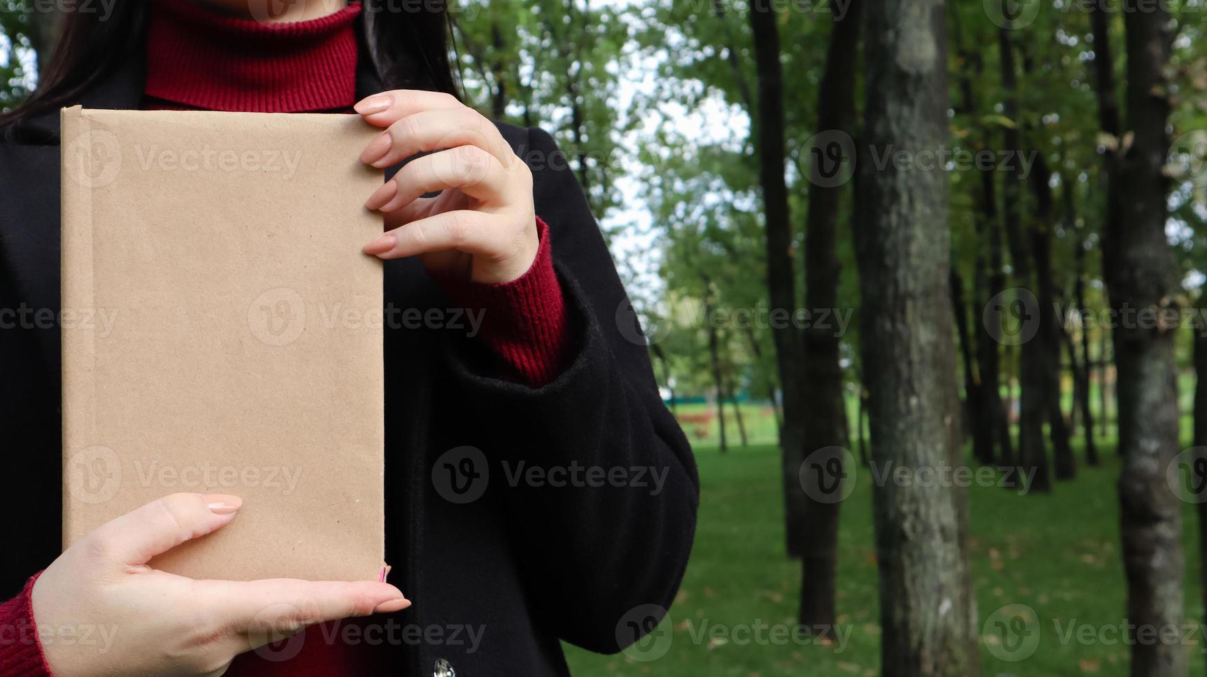 Close up view of woman in black woolen coat and burgundy sweater holding a book with empty cover in the park. Free space for your book reading concept background layout. photo