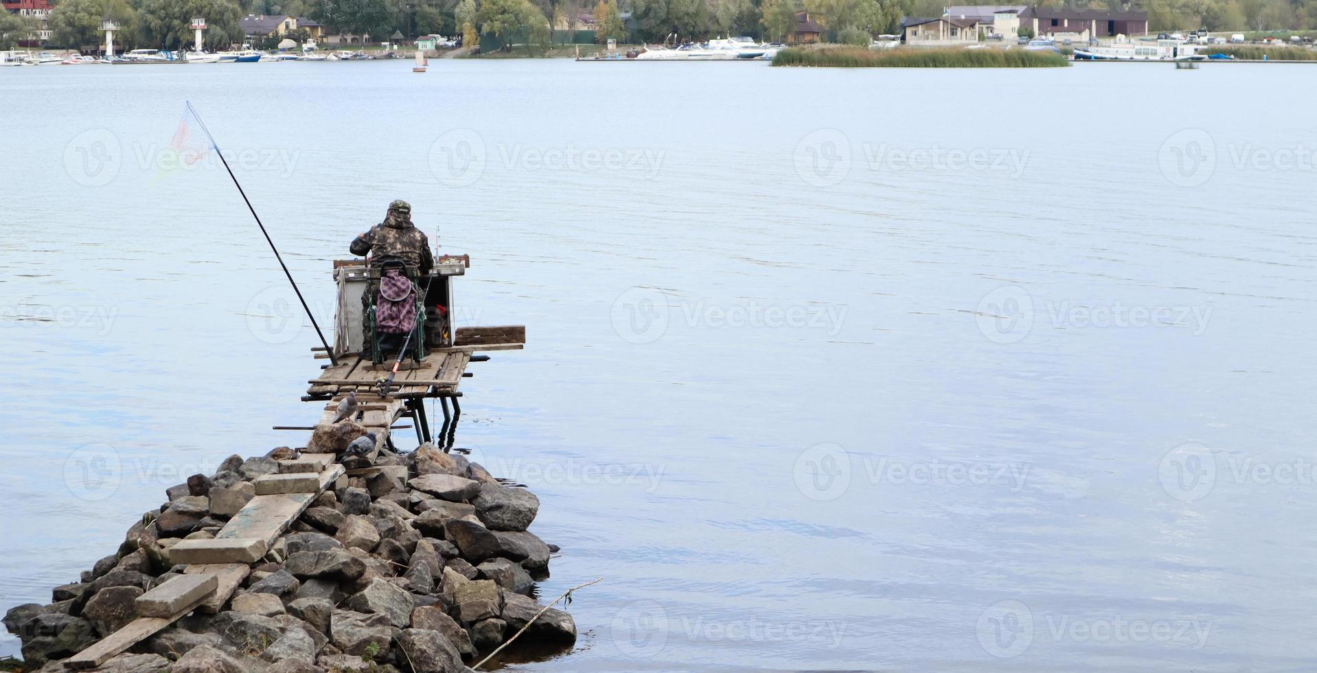 A fisherman catches fish on the river, rear view, from the bank. A fisherman sits on a wooden and stone bridge on the river bank and tries to catch fish. Sports, recreation, lifestyle photo