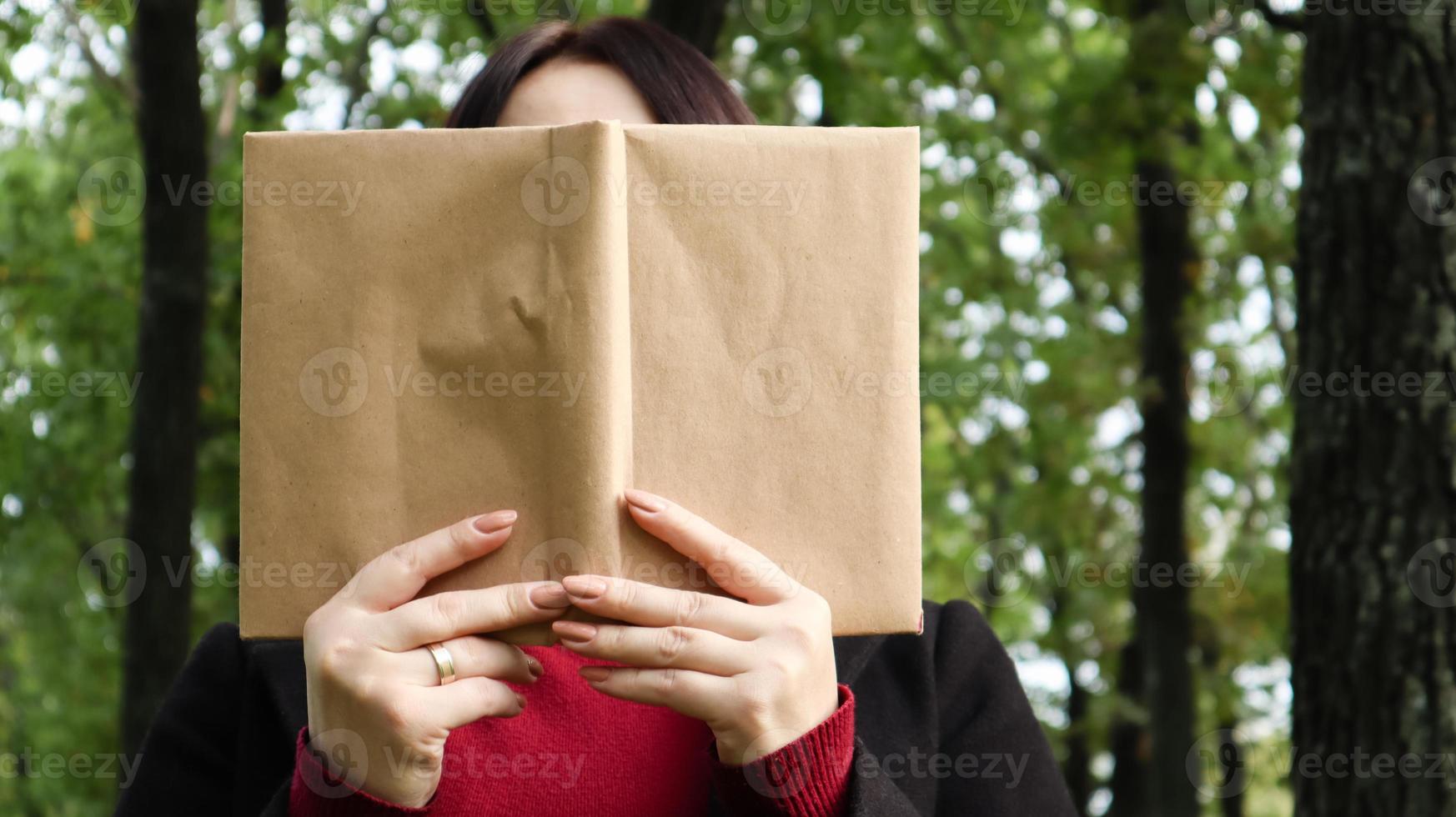 Portrait of a young woman with an open untitled book held close to her face. Brunette covering her face with a book in the park on a sunny day. Education and people concept, selective focus. photo