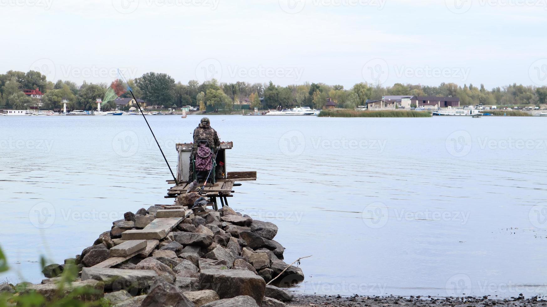 A fisherman catches fish on the river, rear view, from the bank. A fisherman sits on a wooden and stone bridge on the river bank and tries to catch fish. Sports, recreation, lifestyle photo