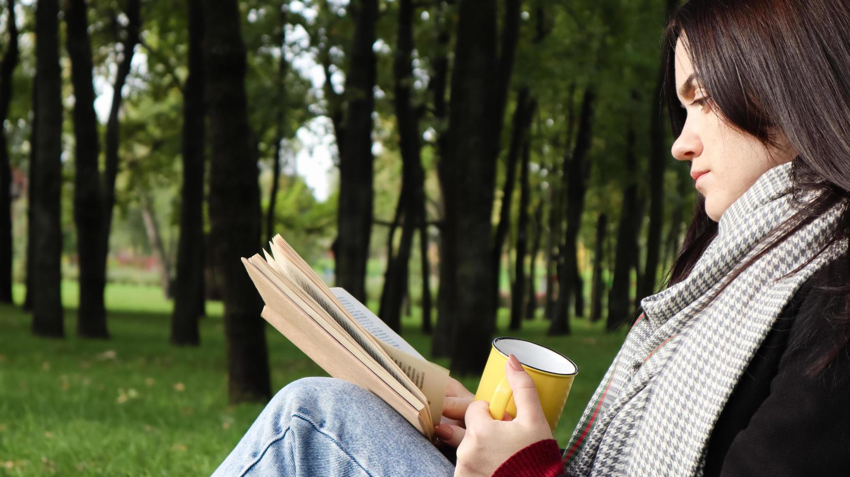 una mujer se sienta debajo de un árbol y lee su libro favorito mientras bebe café o té de una taza amarilla en un parque de la ciudad sobre hierba verde en un agradable día soleado. concepto de vacaciones, educación y estudio. foto