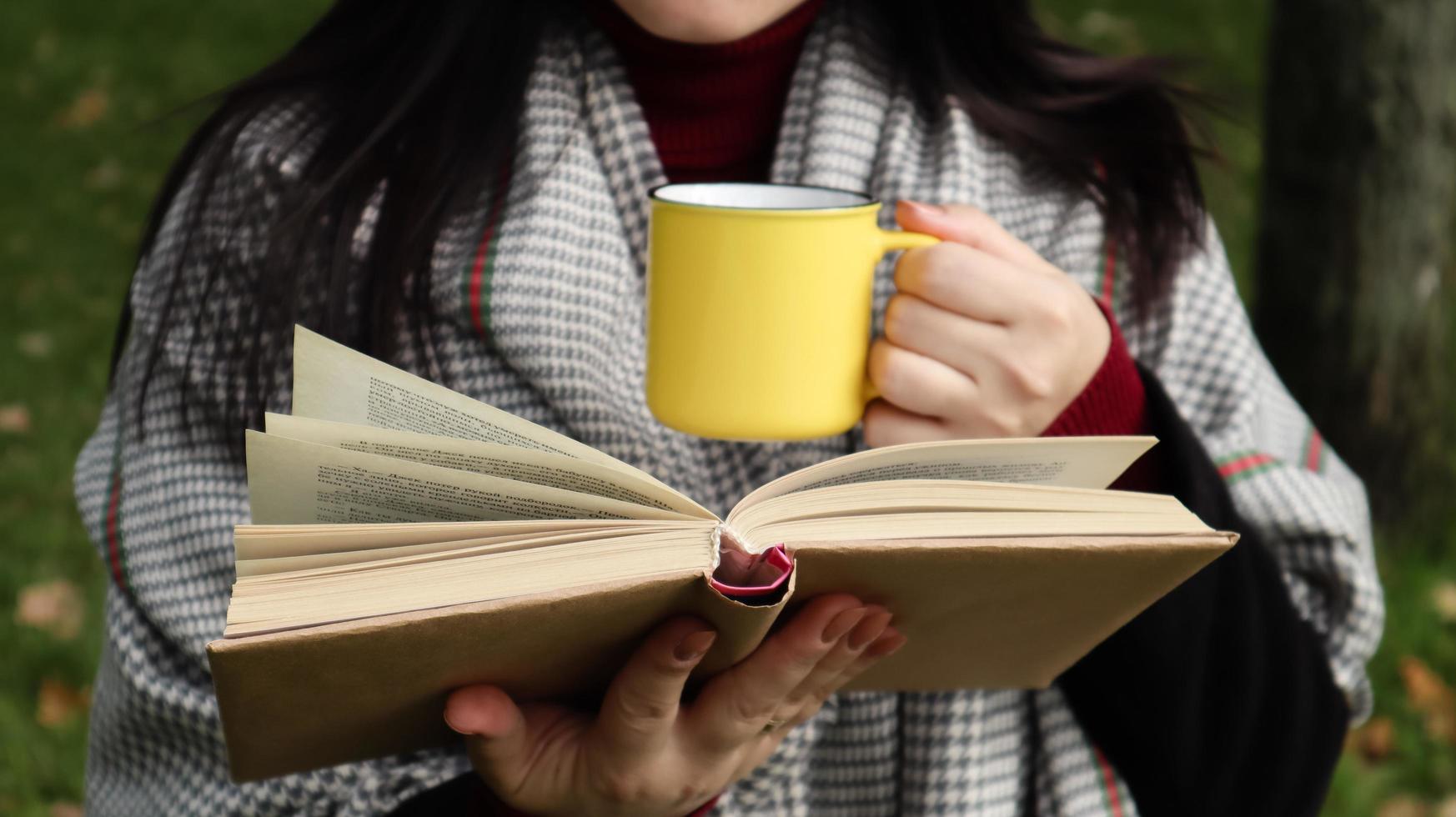 A girl dressed in a coat and a scarf in the autumn forest holds a book and a cup with a hot drink in her hands close-up in a city park on a warm day. The concept of reading, relaxation and comfort. photo