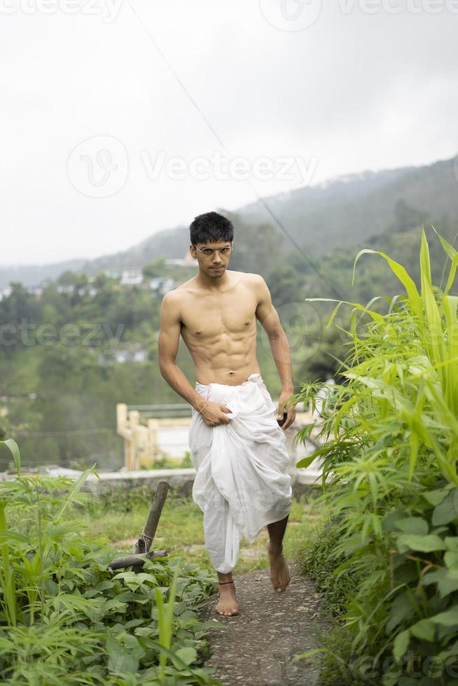 joven indio en forma, caminando por un camino al lado de los cultivos en el campo. un sacerdote indio caminando vestido con un dhoti blanco. hombre religioso indio. foto