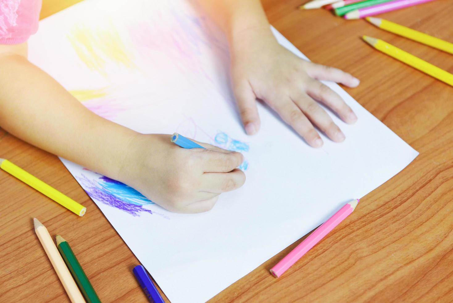 Niña pintando sobre una hoja de papel con lápices de colores sobre la mesa de madera en casa - niño chico haciendo dibujos y lápices de colores foto