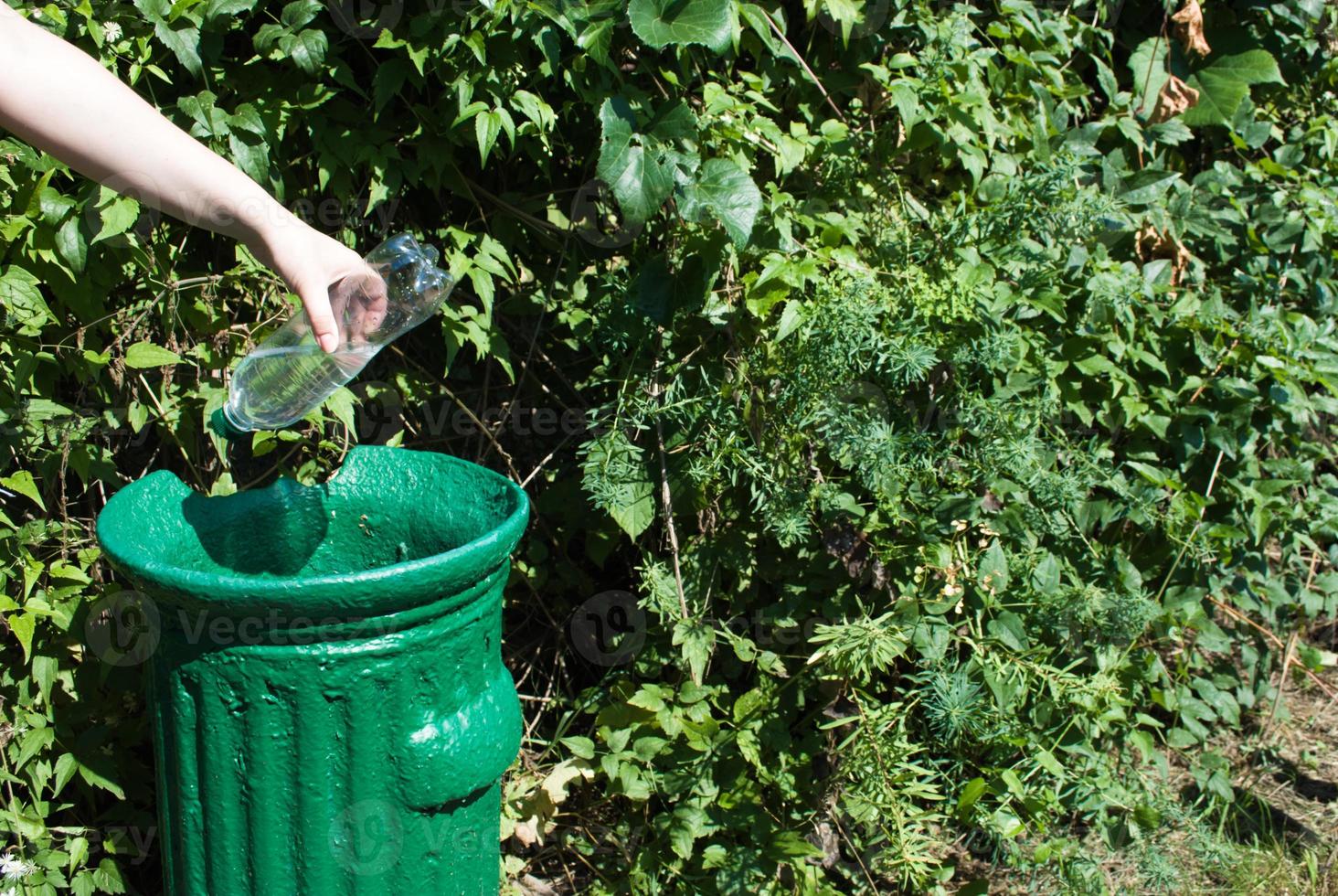 Female hand throws a plastic bottle into a small iron trash can in a green park in summer, close up. nature protection from plastic. photo