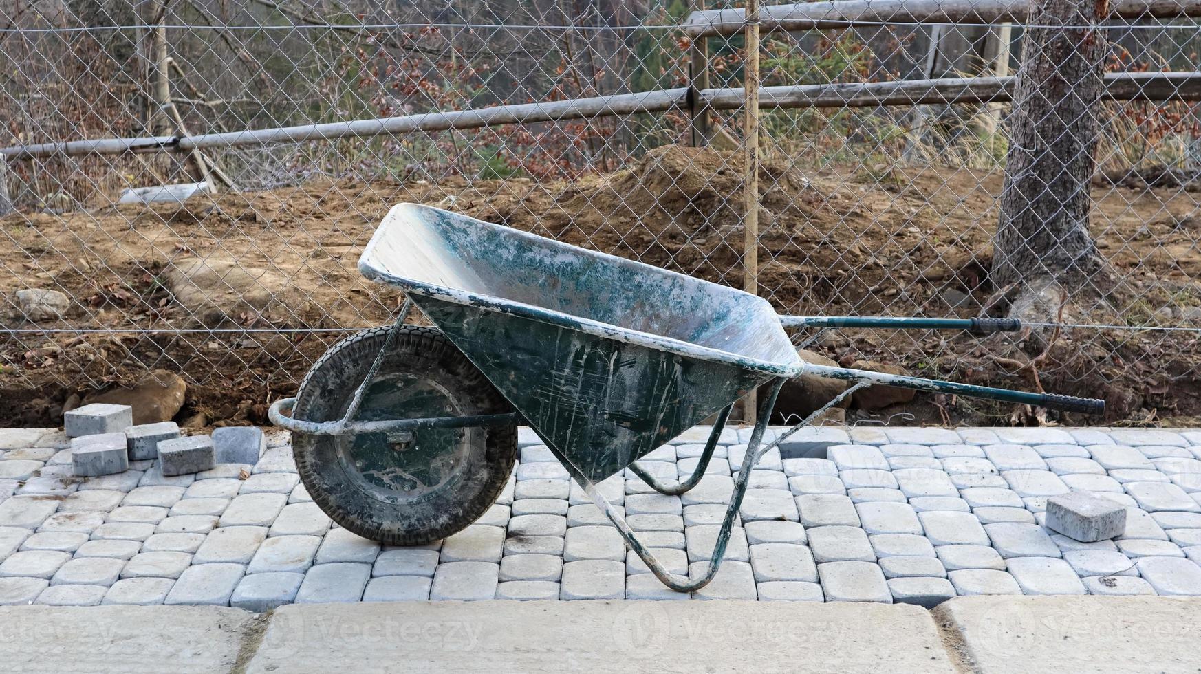 An old green metallic hand wheelbarrow with one wheel and handles stands at a construction site. device for transporting goods. Consists of a cargo body, one or more wheels and a handle. photo