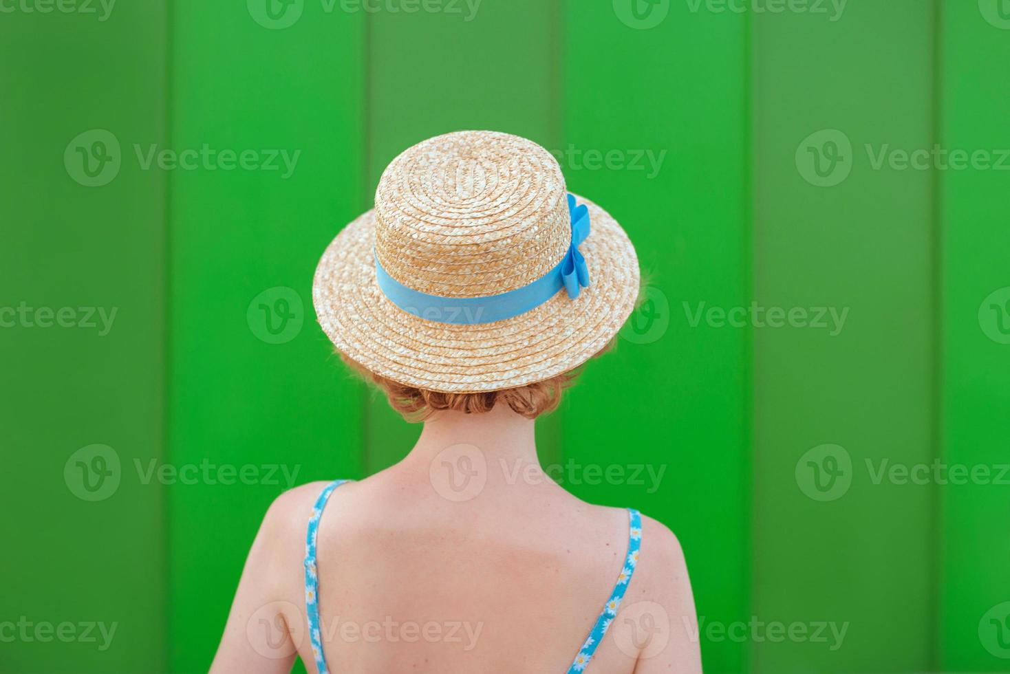 back of young curly redhead woman in straw hat and blue sundress standing on green wall background. Fun, summer, fashion, youth concept. Copy space photo