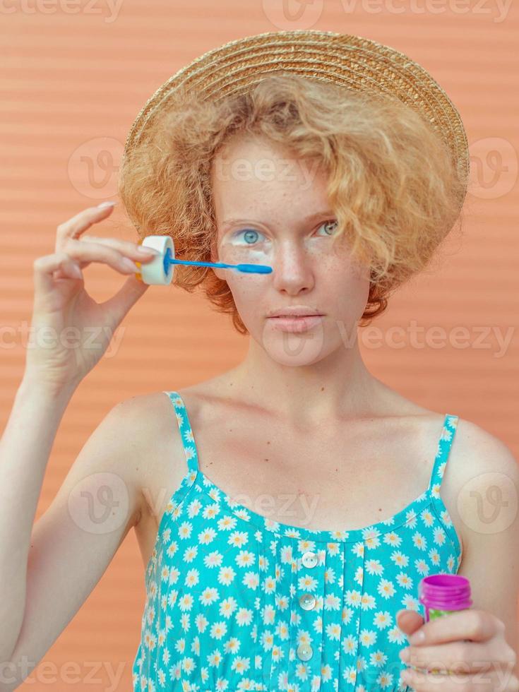 young cheerful curly redhead woman in blue sundress and straw hat with bubbles on beige background. Fun, summer, fashion, youth concept photo