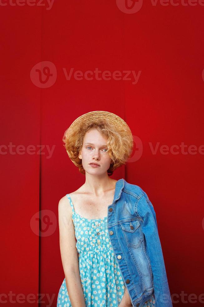 young curly redhead woman in straw hat, blue sundress and jeans jacket standing on dark red background. Fun, summer, fashion, youth concept. Copy space photo