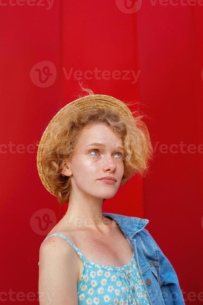 young curly redhead woman in straw hat, blue sundress and jeans jacket standing on dark red background. Fun, summer, fashion, youth concept. Copy space photo