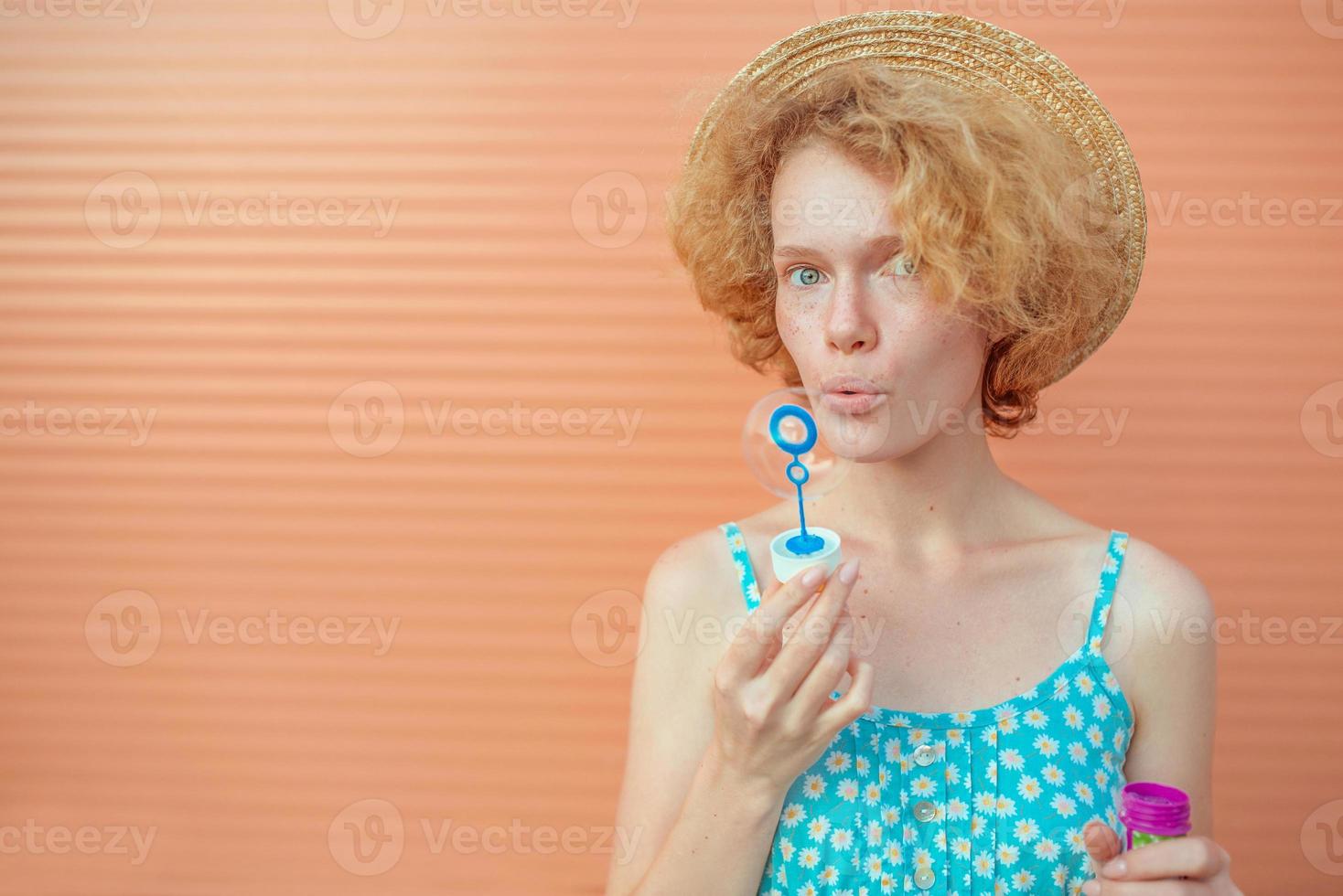 young cheerful curly redhead woman in blue sundress and straw hat with bubbles on beige background. Fun, summer, fashion, youth concept photo