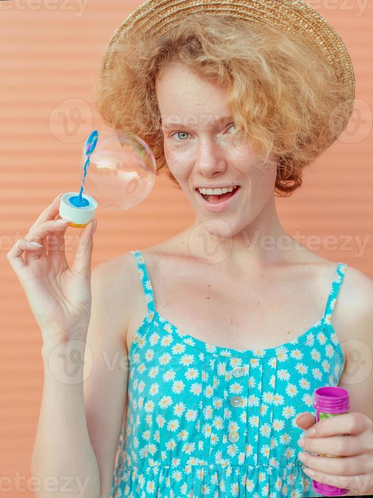 young cheerful curly redhead woman in blue sundress and straw hat with bubbles on beige background. Fun, summer, fashion, youth concept photo