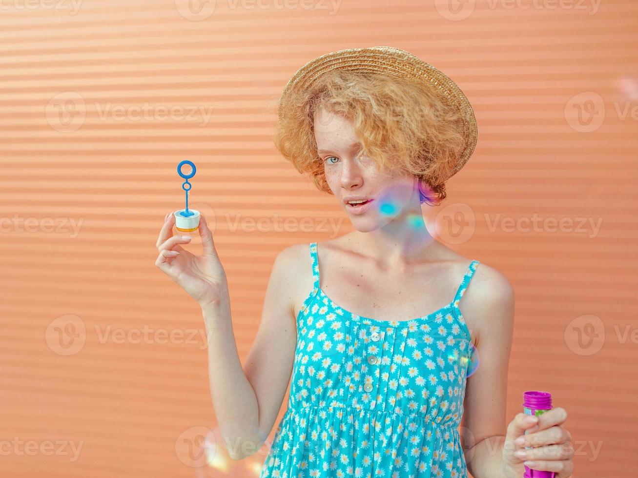 young cheerful curly redhead woman in blue sundress and straw hat with bubbles on beige background. Fun, summer, fashion, youth concept photo