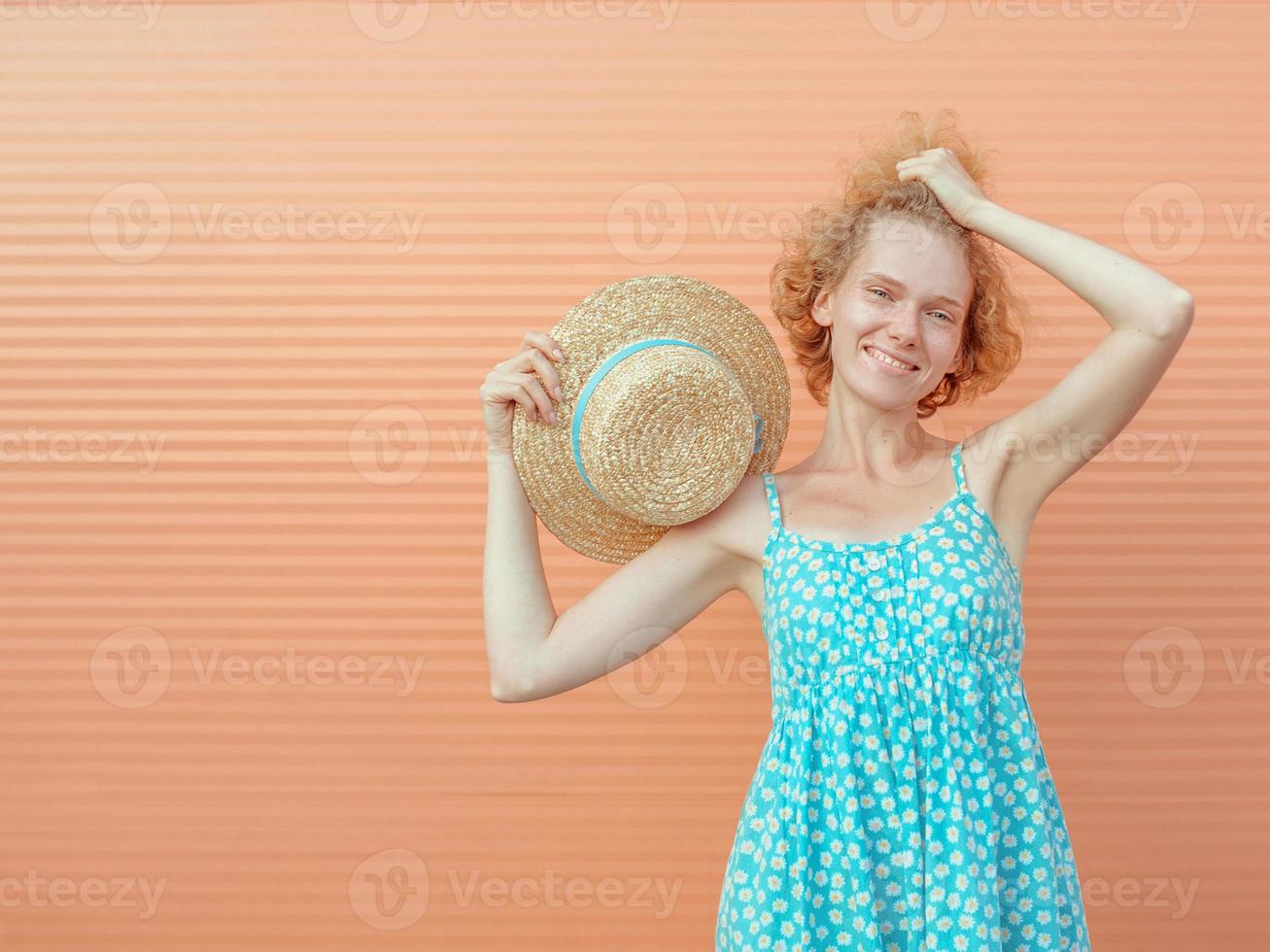 young cheerful curly redhead woman in blue sundress holding straw hat in her hand on beige background. Fun, summer, fashion, youth concept photo