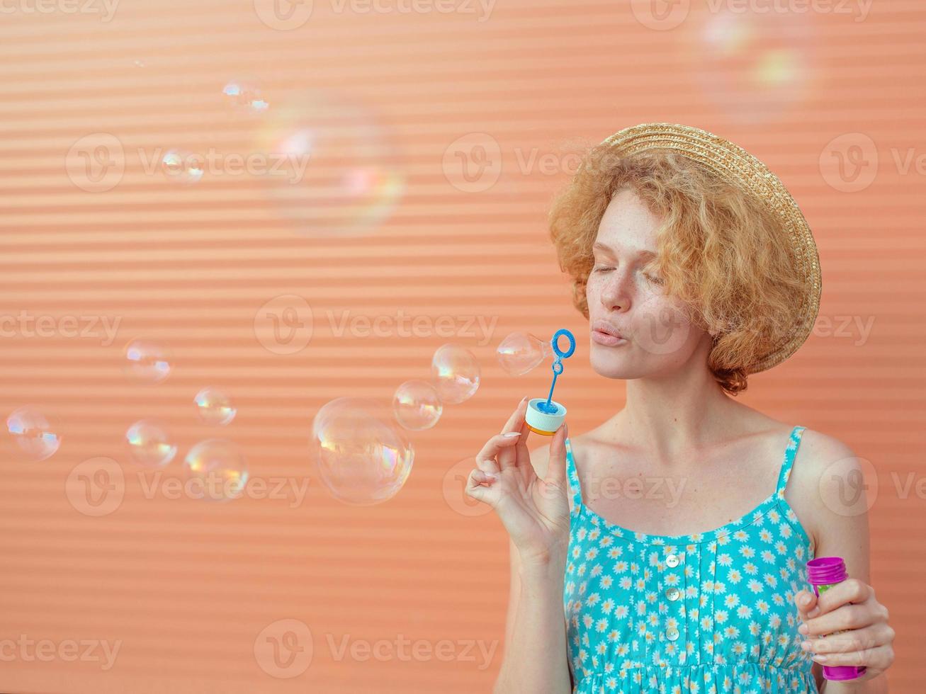 young cheerful curly redhead woman in blue sundress and straw hat with bubbles on beige background. Fun, summer, fashion, youth concept photo